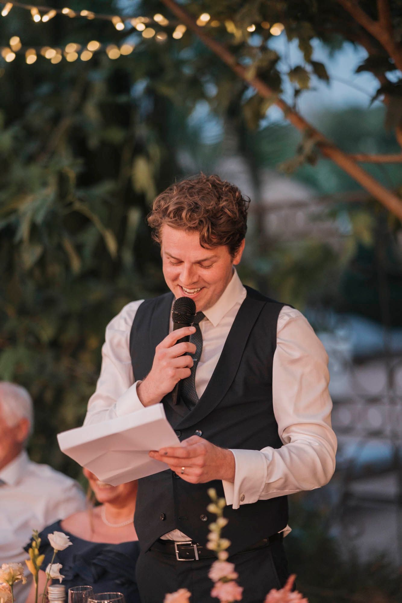 A person in formal attire gives a speech at an outdoor wedding reception under string lights.