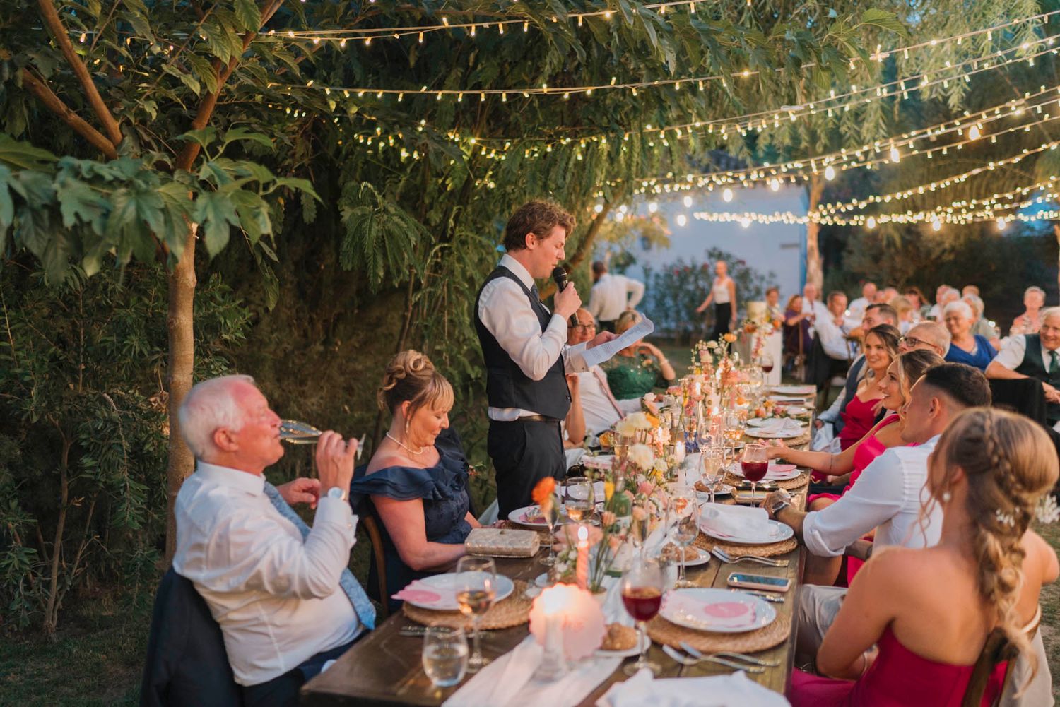 Wedding reception dinner table setup outdoors with string lights and evening ambiance.