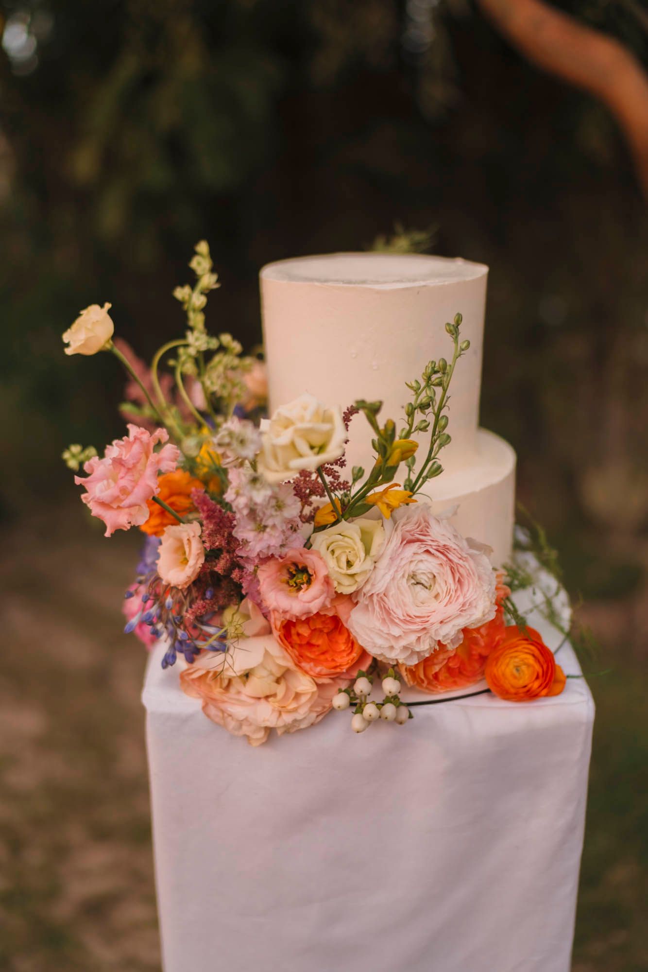 An elegant white wedding cake decorated with fresh pink, orange and purple flowers against a dark background.