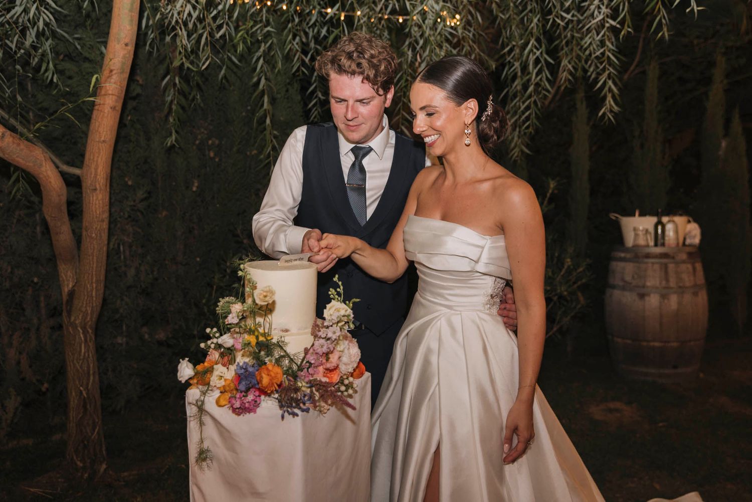 A couple stands next to their floral-adorned wedding cake at an evening outdoor reception.