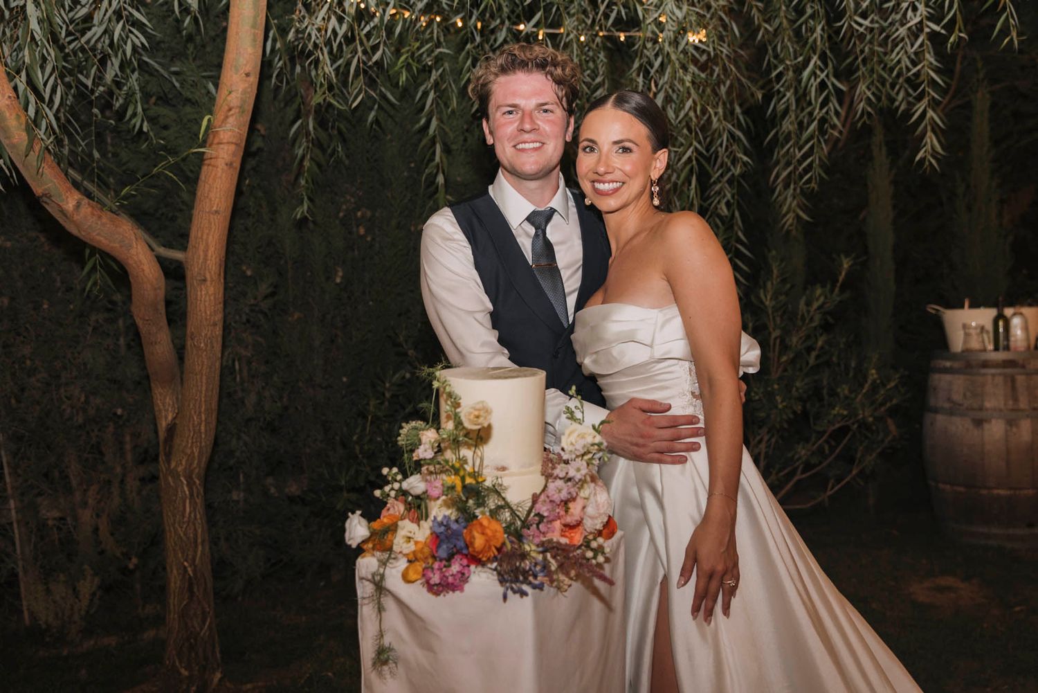 A happy couple poses with their elegant wedding cake under willow trees at dusk.