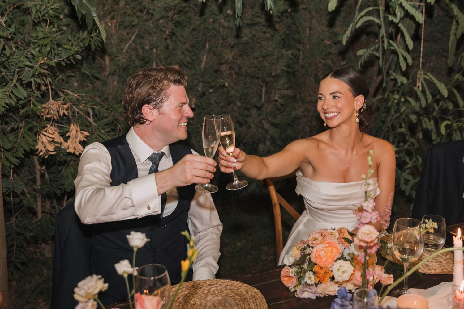 A couple toasts with champagne glasses at their outdoor reception table.