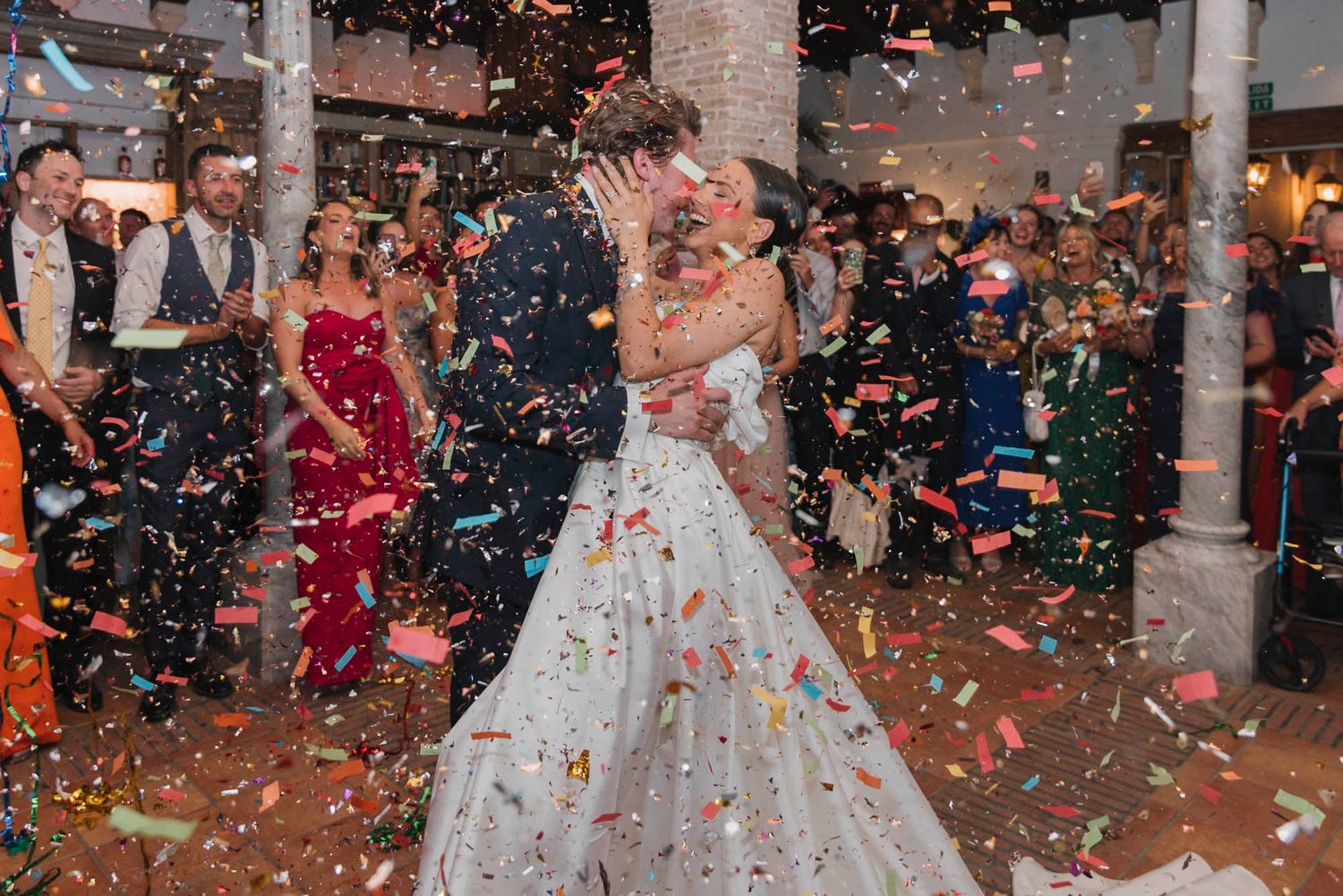 A couple dances as colorful confetti rains down in an elegant columned venue.