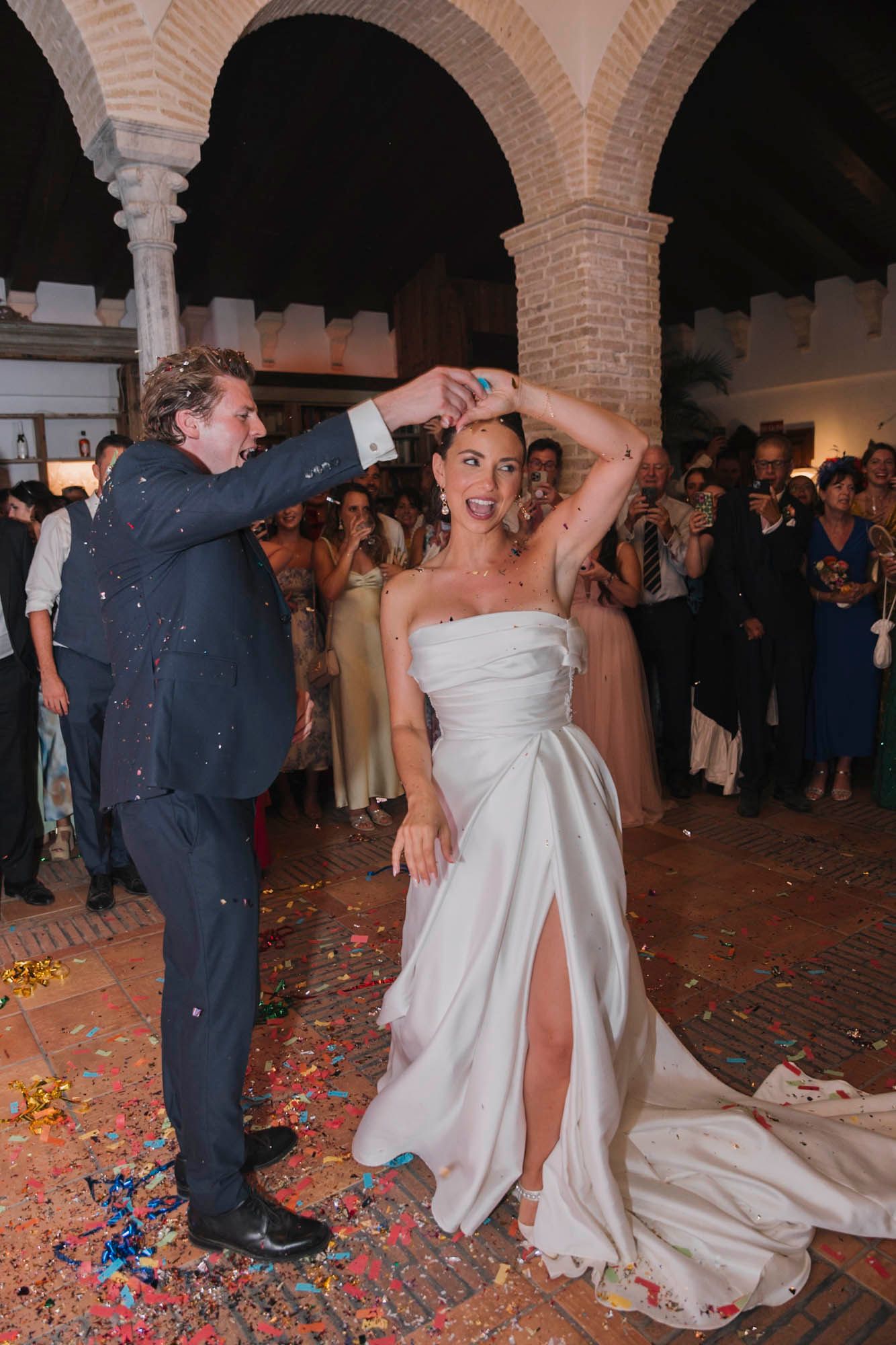 A dancing couple celebrates with confetti falling around them in an arched reception venue.