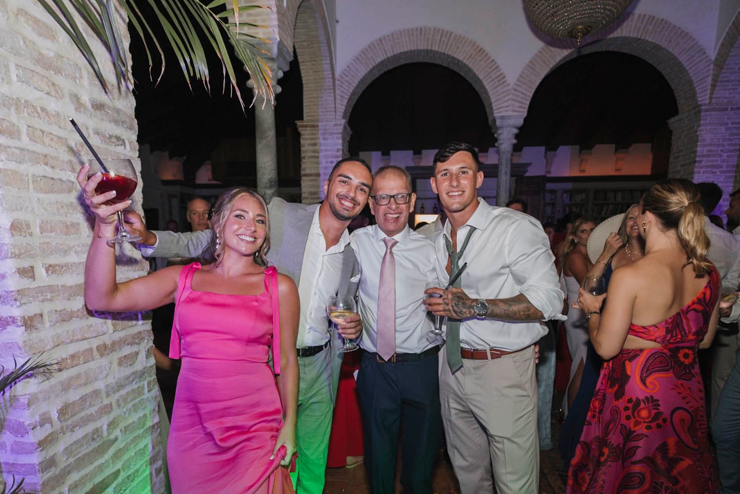 Wedding guests dancing and celebrating at an elegant venue with arched architecture and colorful lighting.