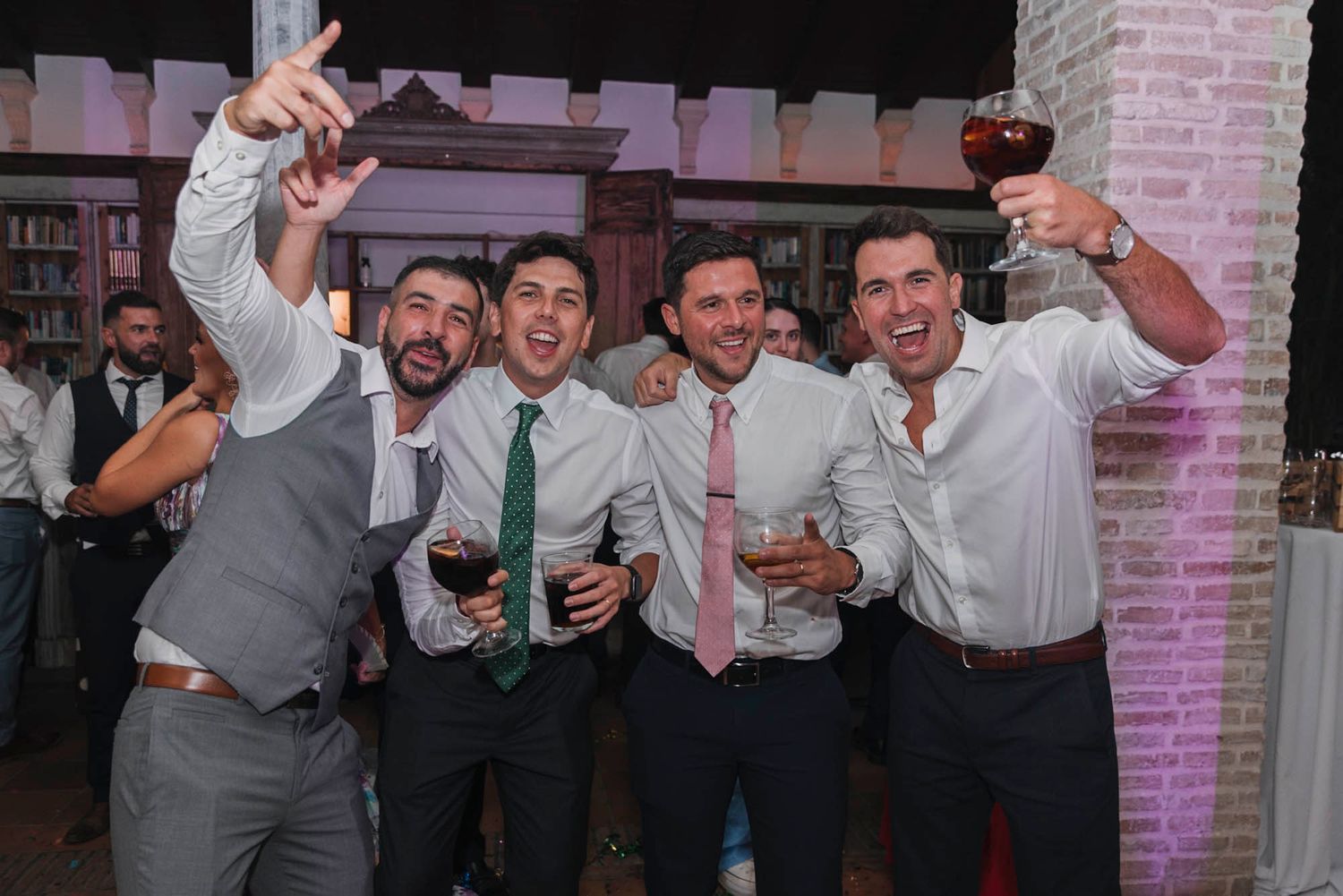 Party guests in formal attire raise their drinks and celebrate at an elegant venue with brick archways.