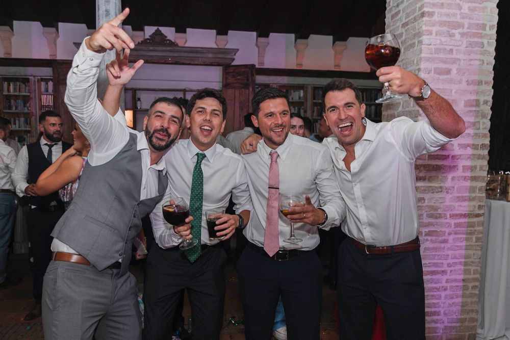 Party guests in formal attire raise their drinks and celebrate at an elegant venue with brick archways.