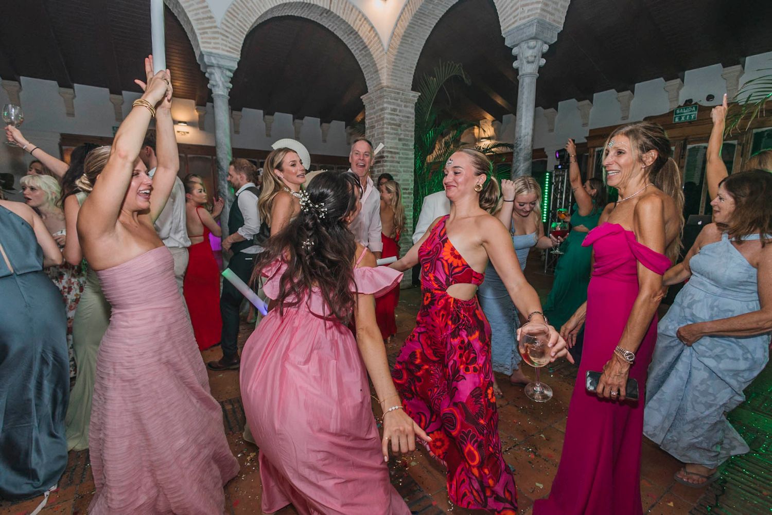 Wedding guests in colorful dresses dancing enthusiastically under ornate arches during reception.