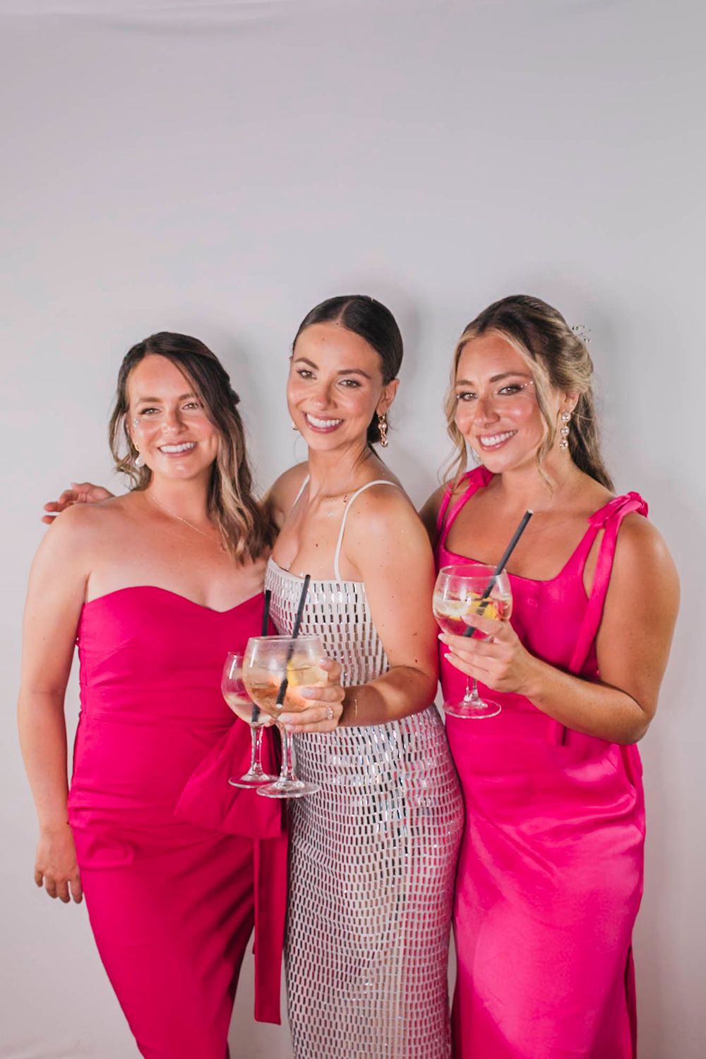 Three women in elegant pink and white dresses pose together holding wine glasses.