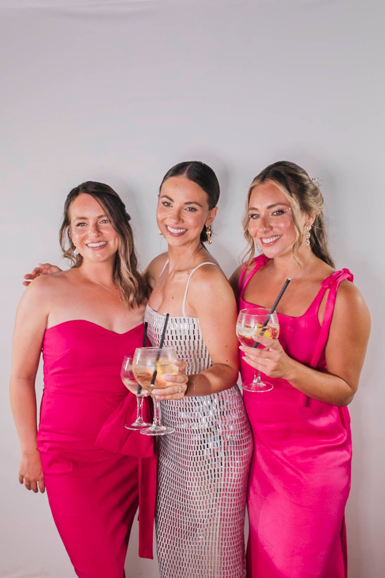Three women in elegant pink and white dresses pose together holding wine glasses.