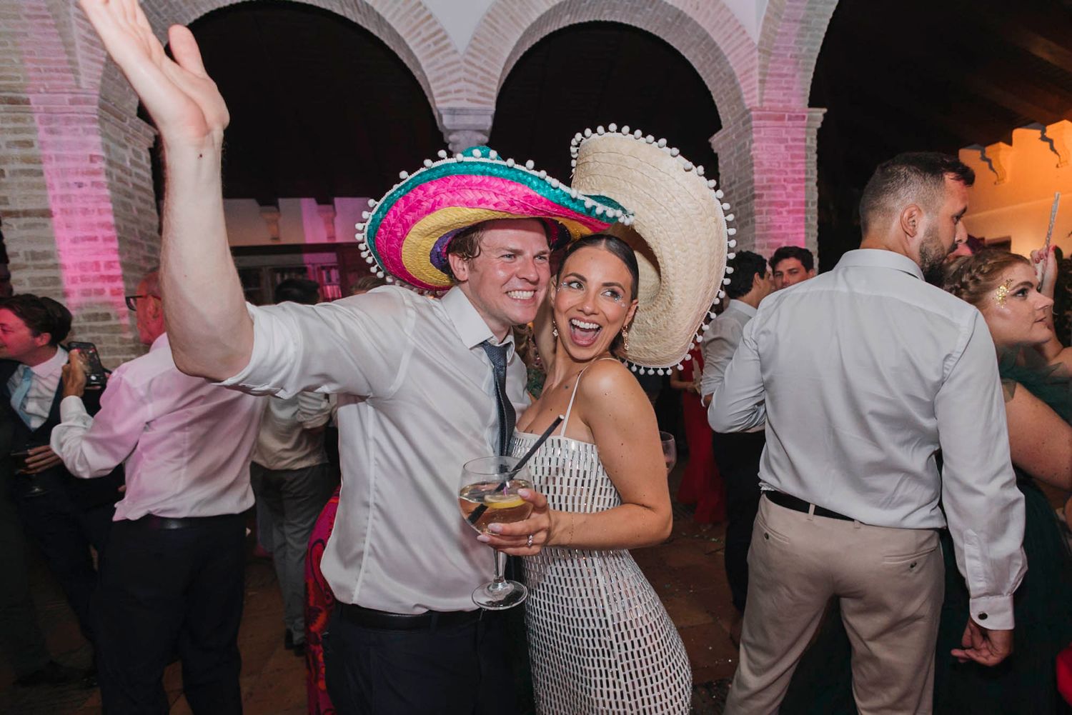 Party guests celebrate wearing festive sombreros under arched brick architecture with mood lighting.
