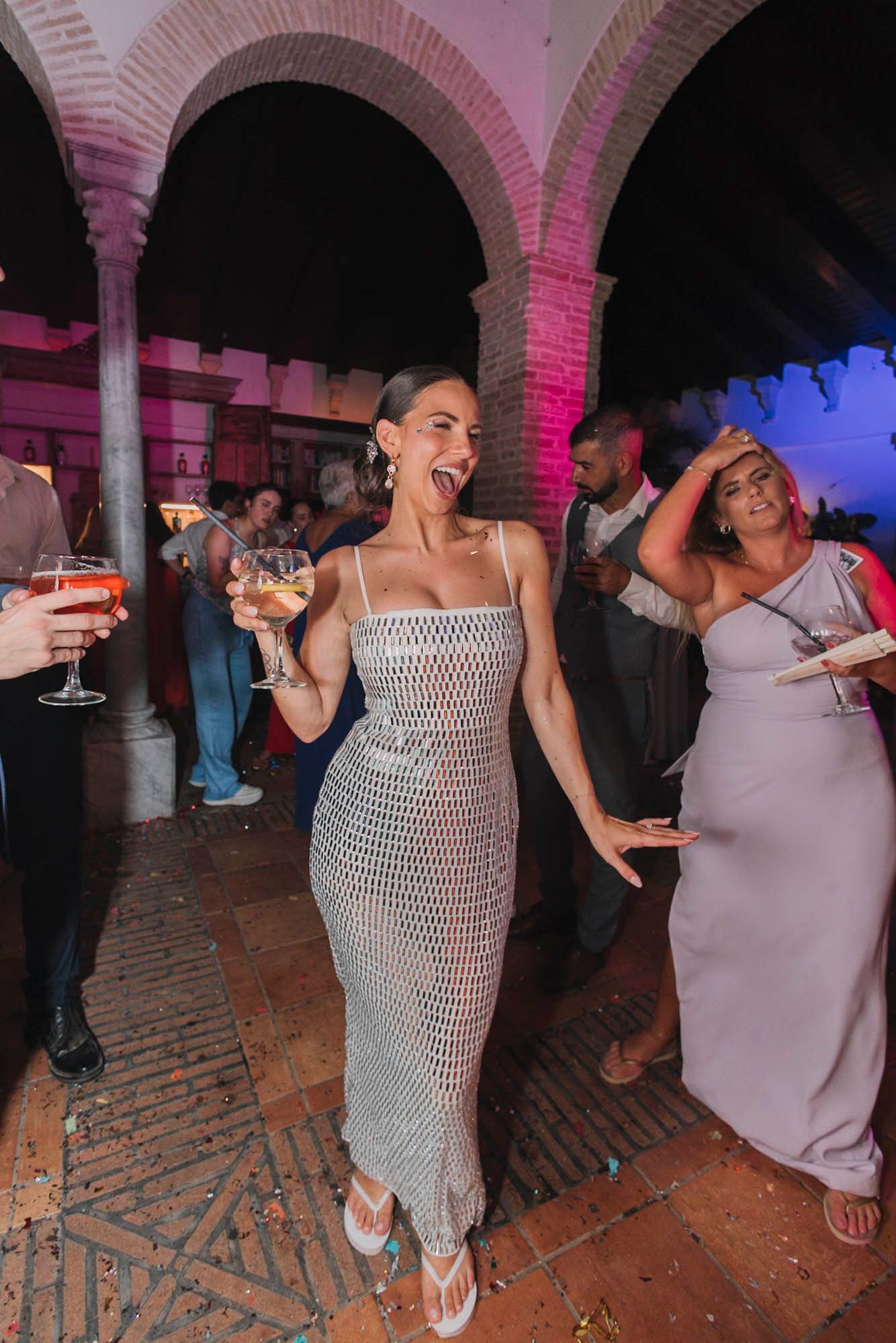 Guest in sparkly dress dances at reception venue with brick arches and colored lighting.