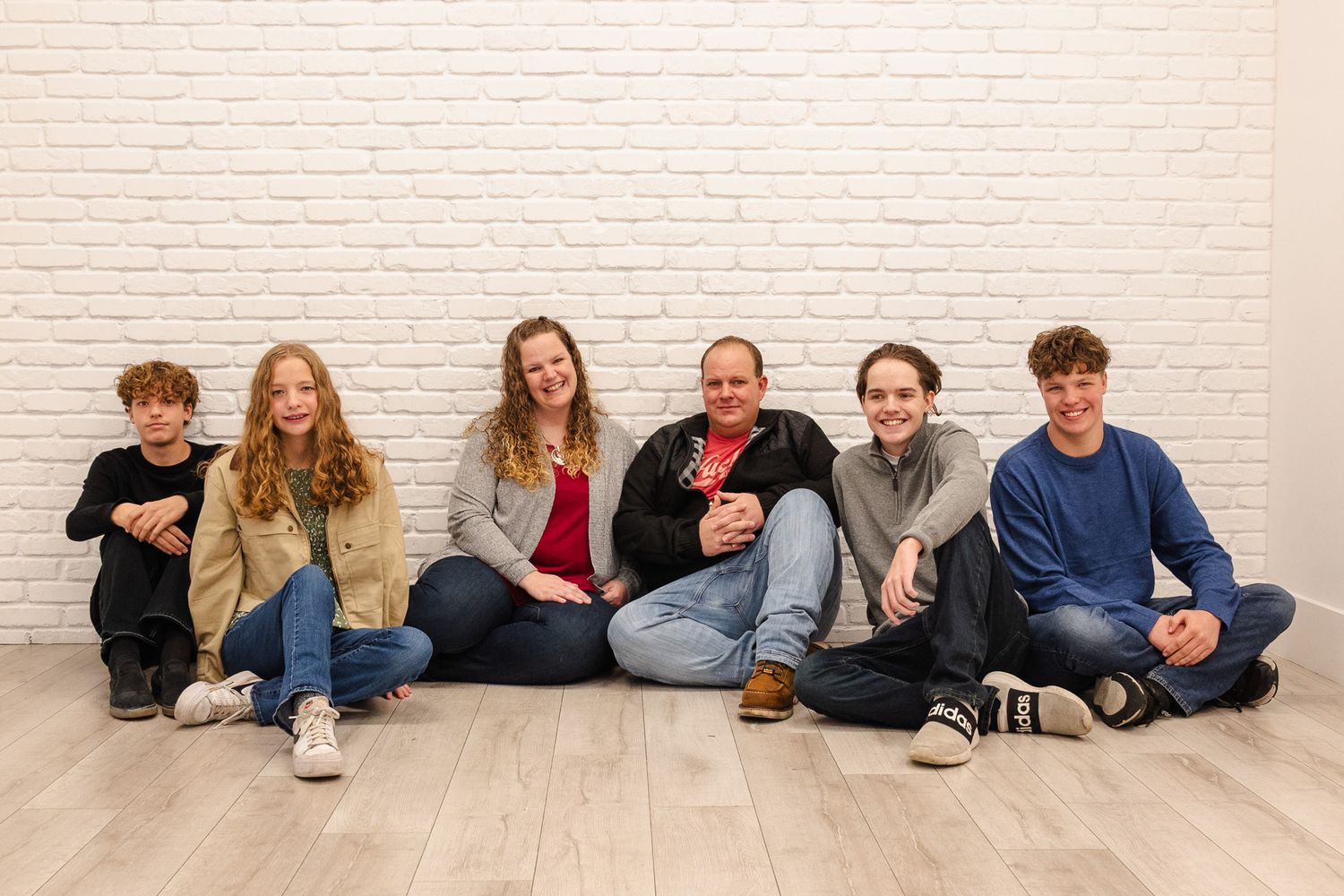 Family of six sitting on floor together smiling in front of white brick wall.