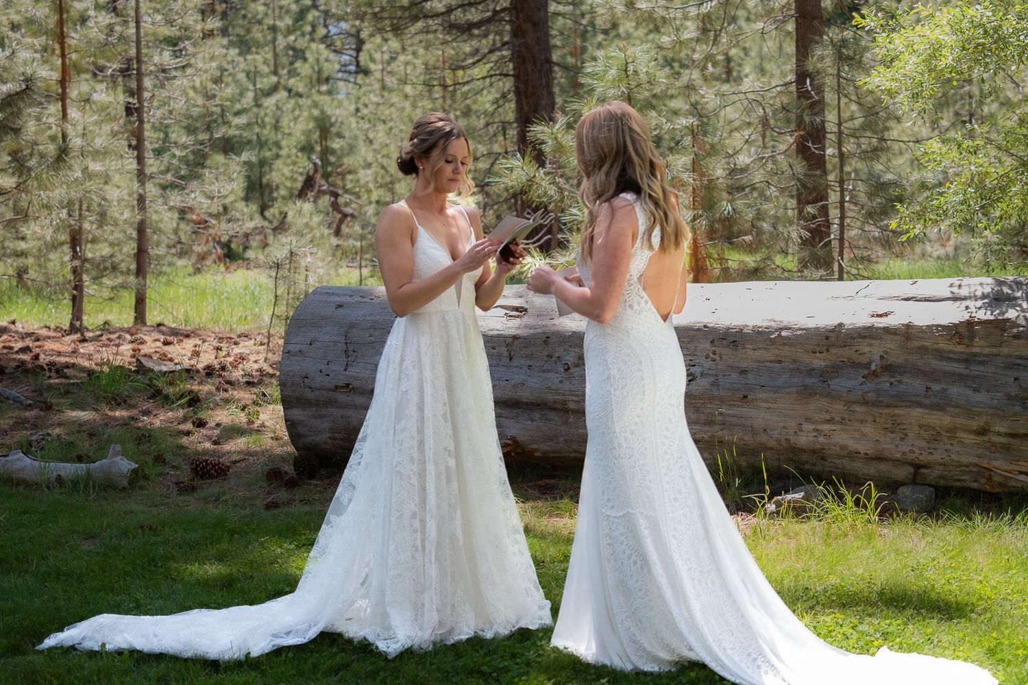 Two brides in elegant white lace gowns share a joyful moment together in a sunlit forest setting.