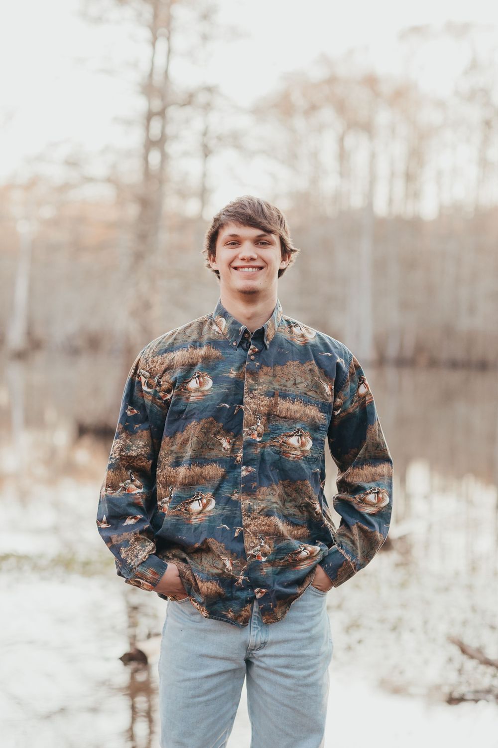 A person wearing a camouflage patterned button-up shirt and light jeans poses outdoors in winter scenery.