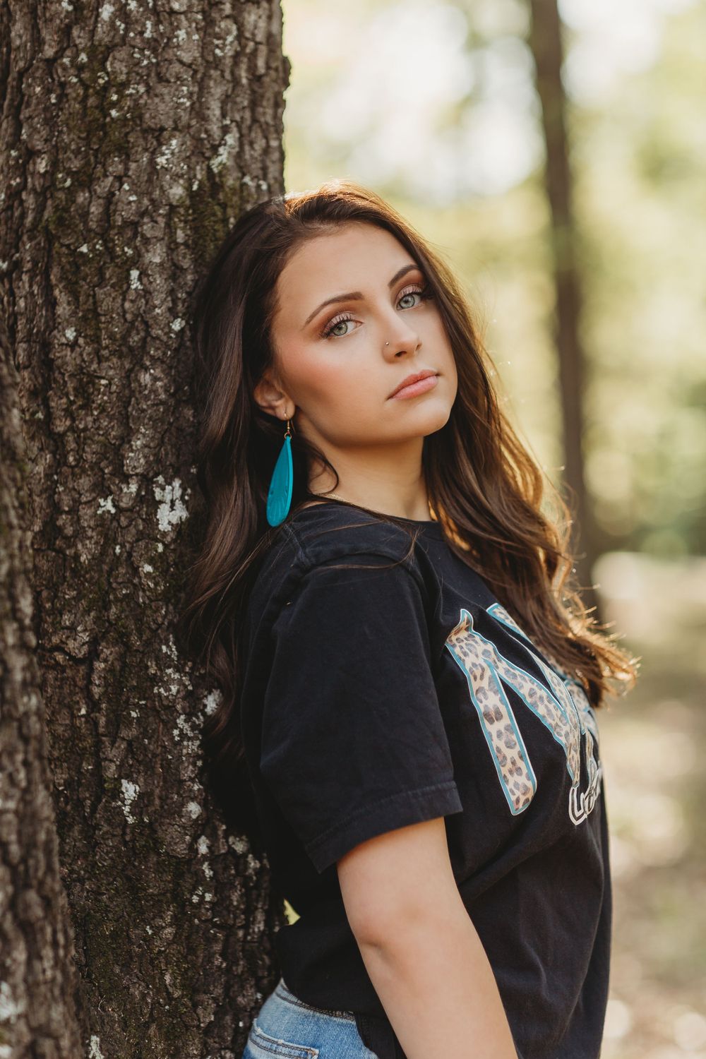 A person in a black t-shirt and turquoise earrings leans against a tree trunk in a sunlit forest setting.
