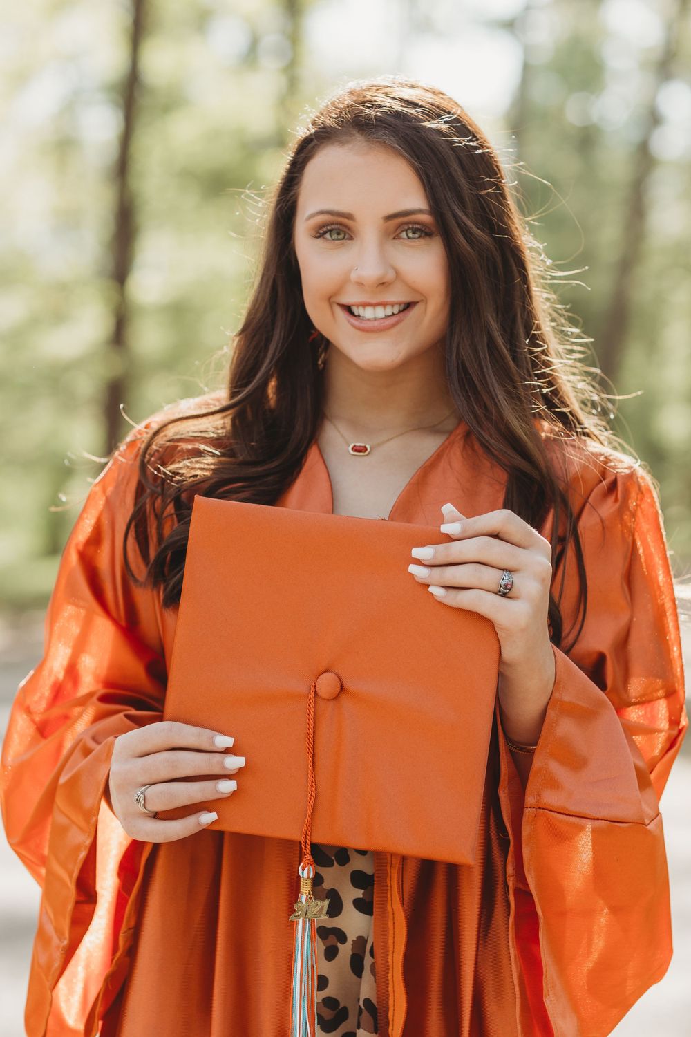Graduate wearing bright orange academic gown holds red diploma folder while smiling outdoors during graduation photo session.