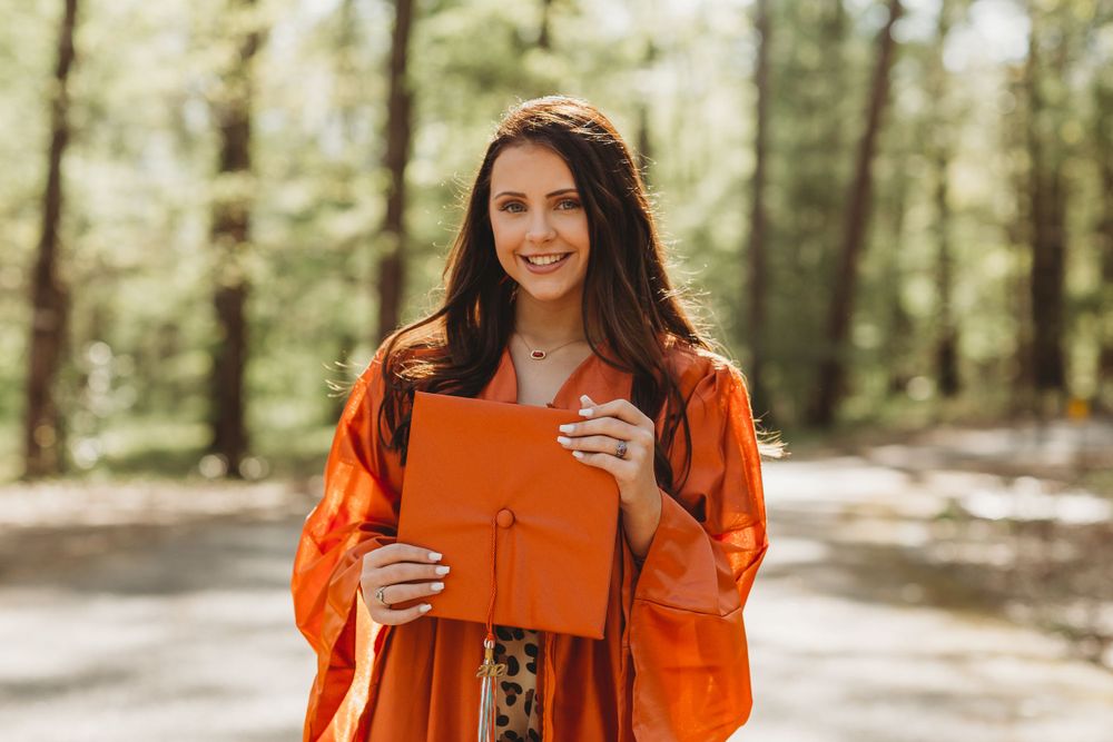 Graduate in bright orange graduation gown holds diploma in sunny outdoor forest setting.