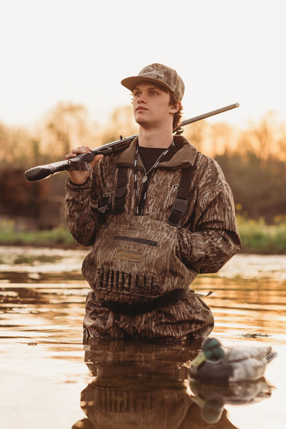 Hunter in camouflage gear stands in water at sunrise with shotgun and duck decoy during waterfowl hunt.