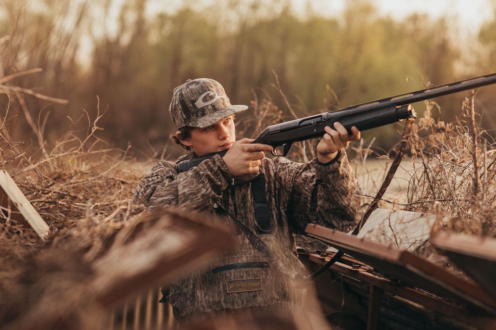 A hunter in camouflage gear aims a shotgun while crouched in dry brush during hunting season.