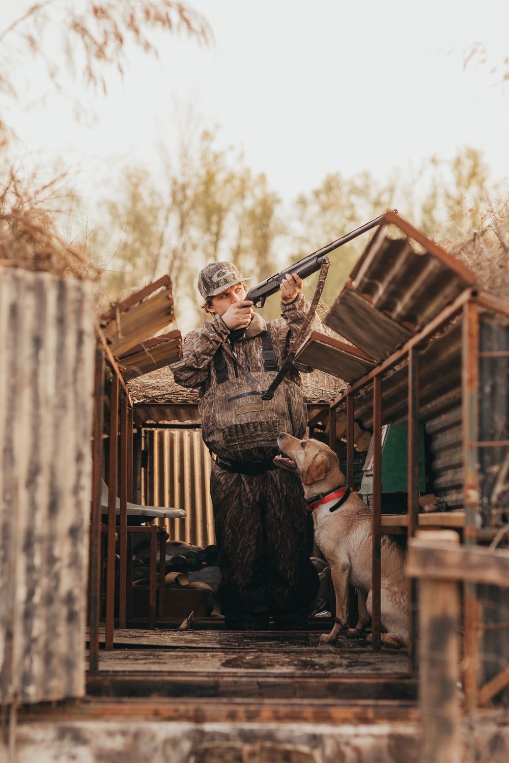 A hunter in camouflage gear stands in a wooden duck blind with their hunting dog during autumn.