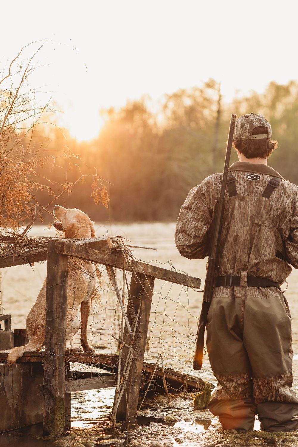 A hunter in camouflage gear and their hunting dog stand on a wooden dock at sunrise during duck hunting season.