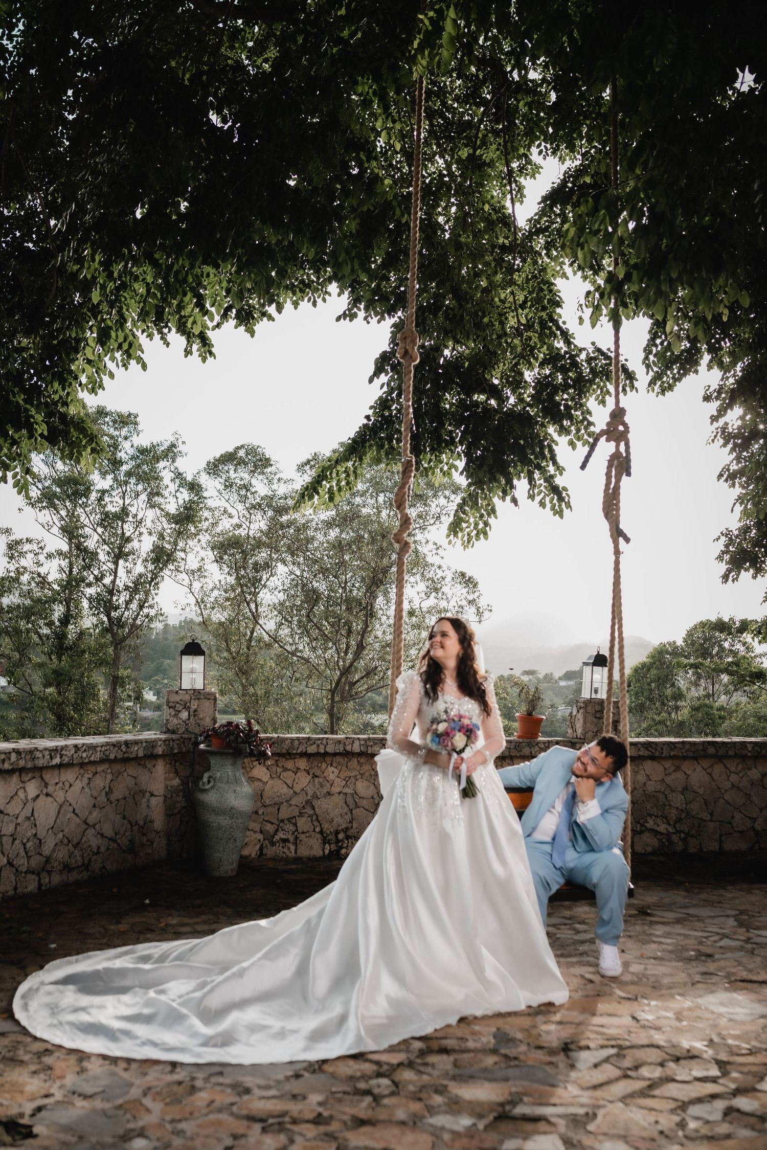 A wedding photo showing a bride in a flowing white gown with a train on a stone terrace surrounded by trees.