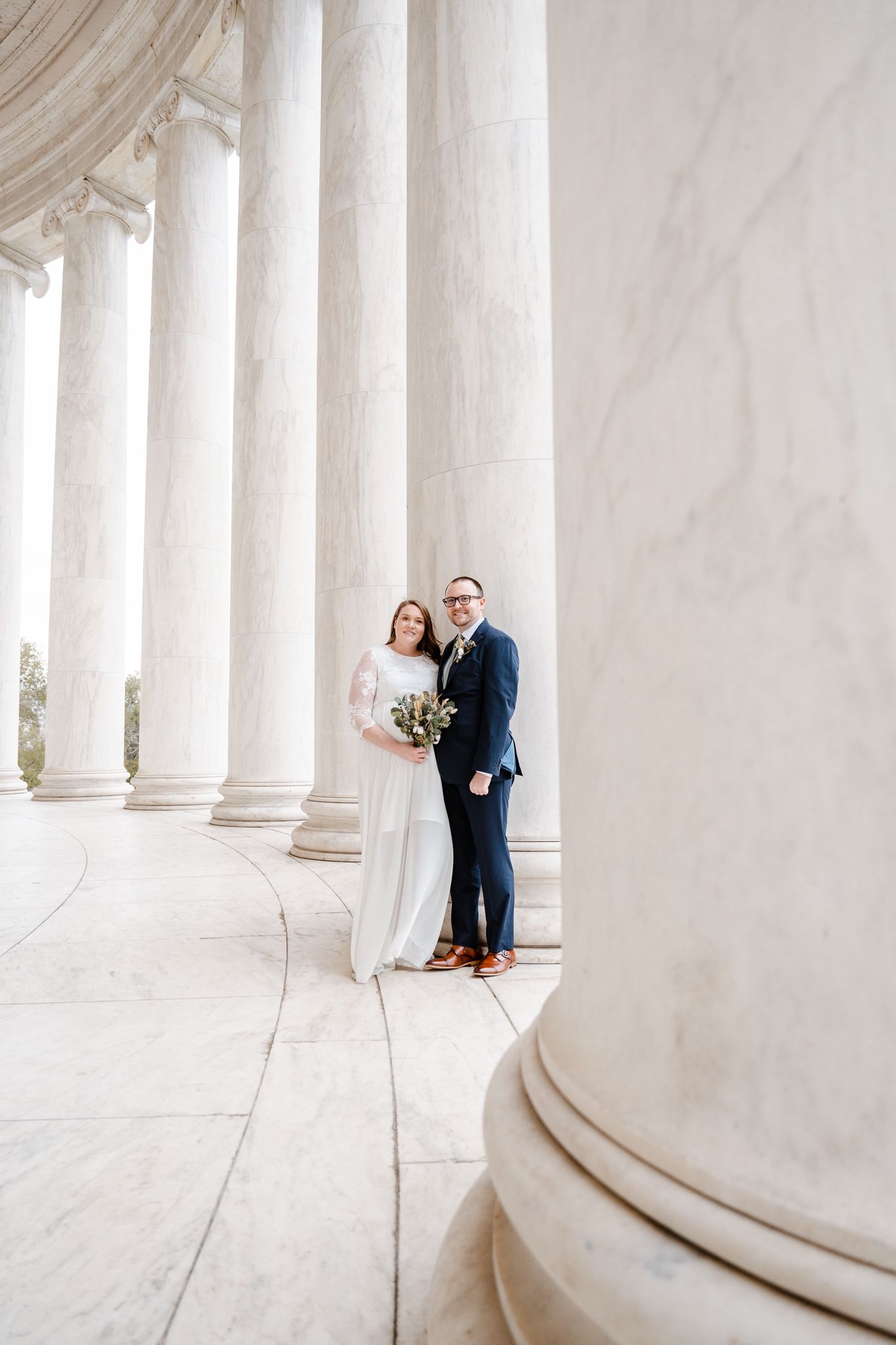 A couple stands among elegant white marble columns at the Jefferson Memorial in Washington DC on their wedding day. Affordable Wedding Photo
