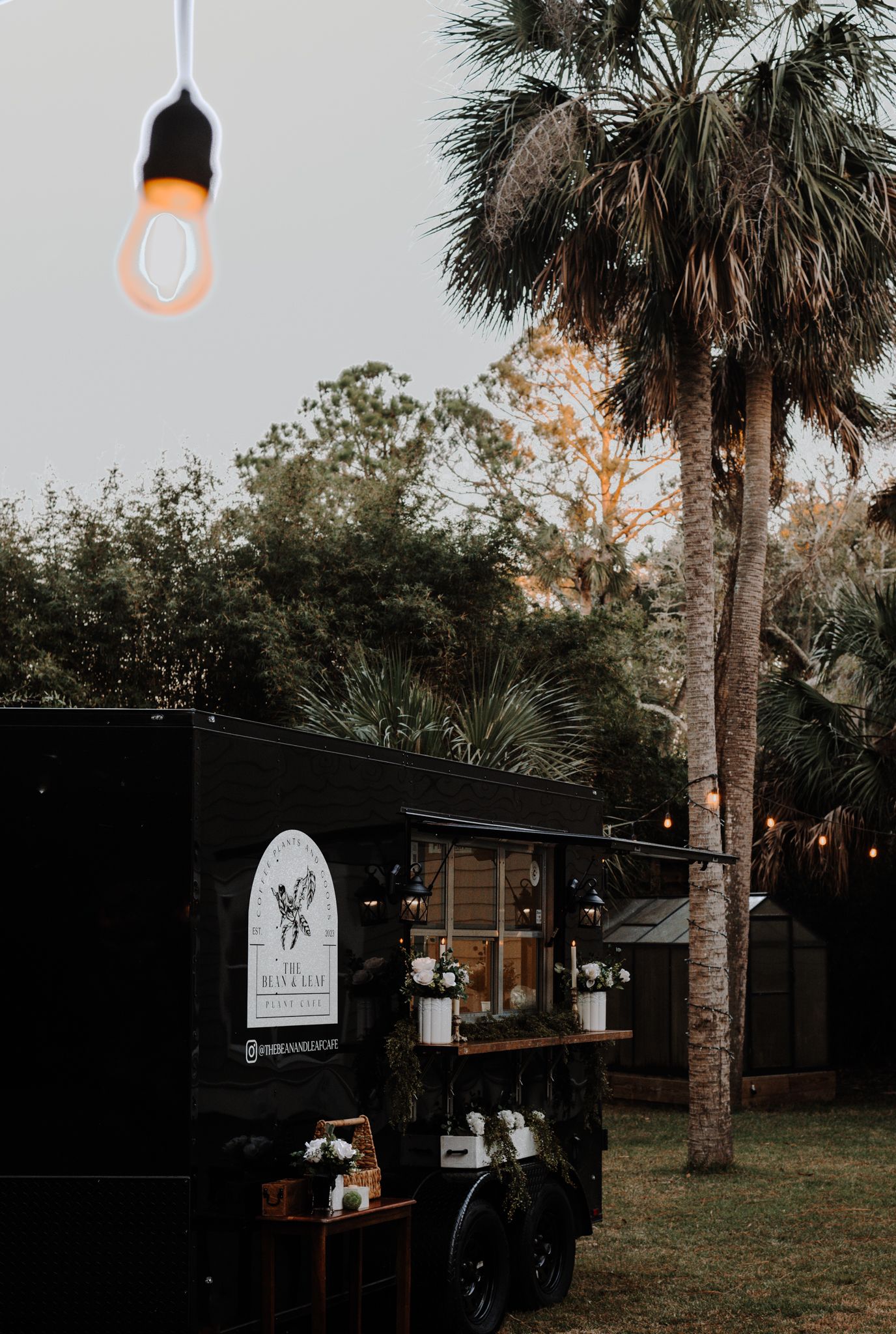 A black coffee truck with hanging lights and palm trees at dusk creates a cozy outdoor ambiance.