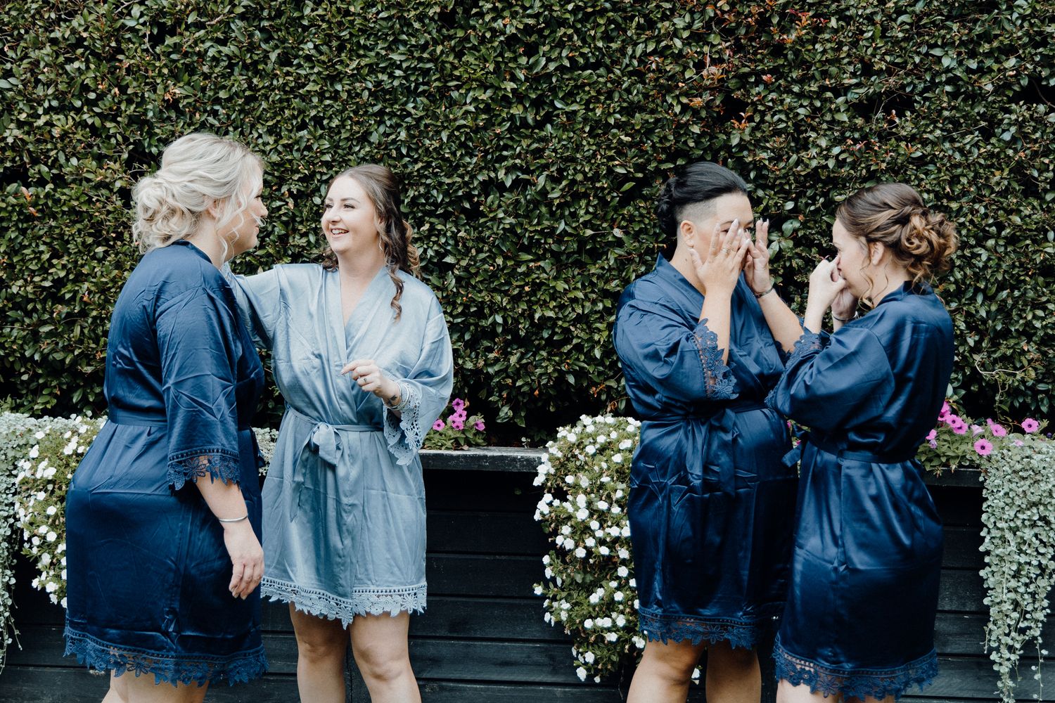Girl Squad in matching blue robes share joyful moments in front of a garden hedge, captured by Apertured Media Auckland Wedding Photographer