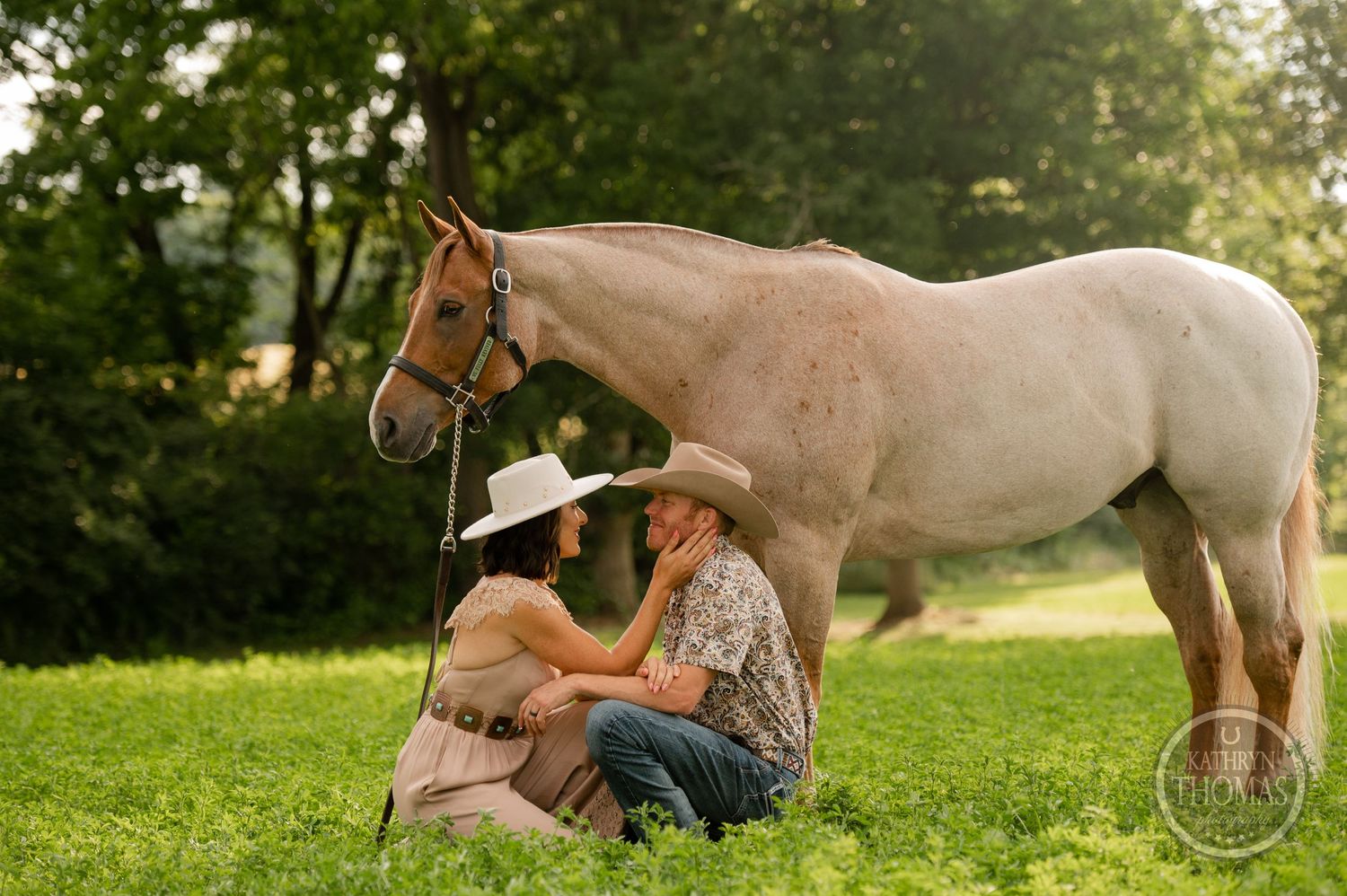 Two people sit in grass beside a cream-colored horse during golden hour.