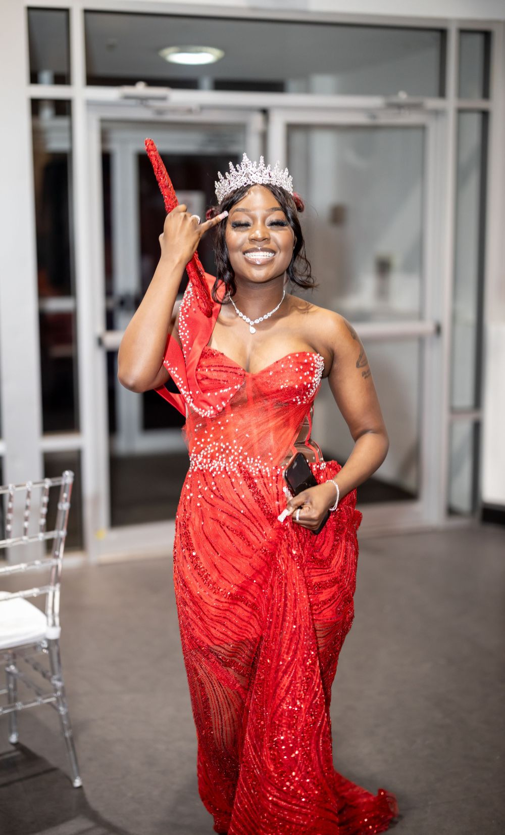 Person in a sparkling red gown and tiara poses at an event entrance.
