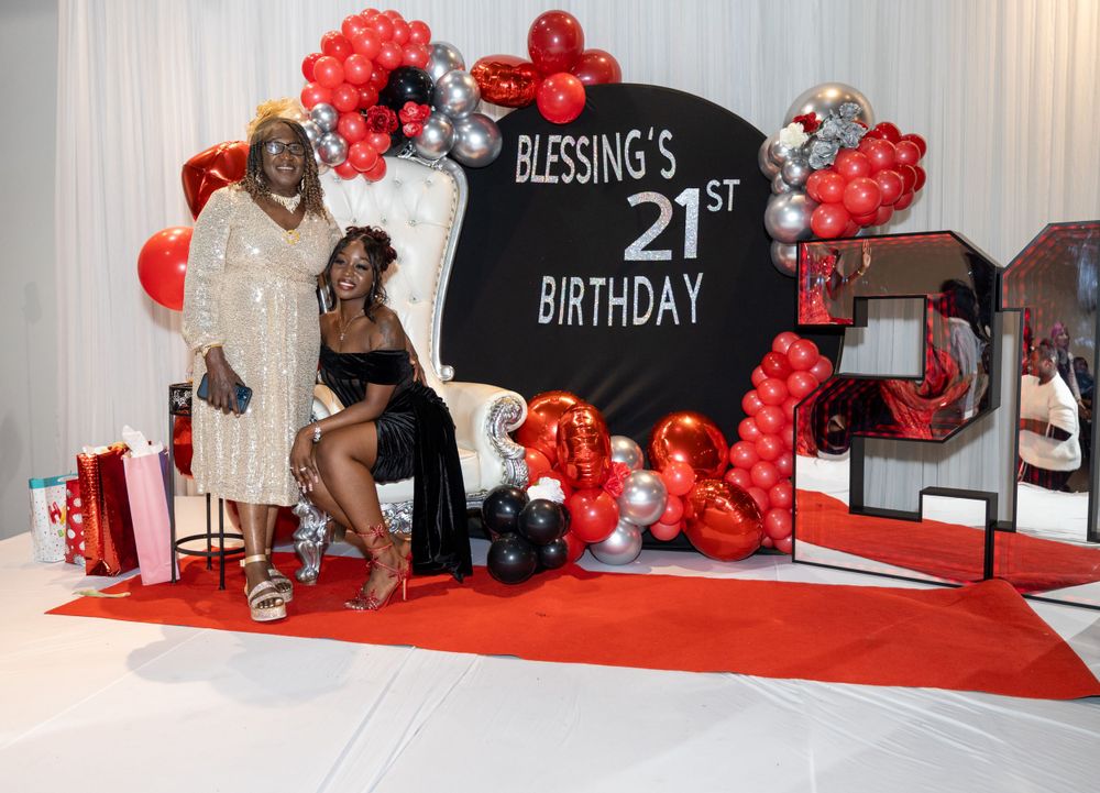Birthday party setup with red and black balloon arch, white throne chair, and decorative '21' numbers on red carpet.