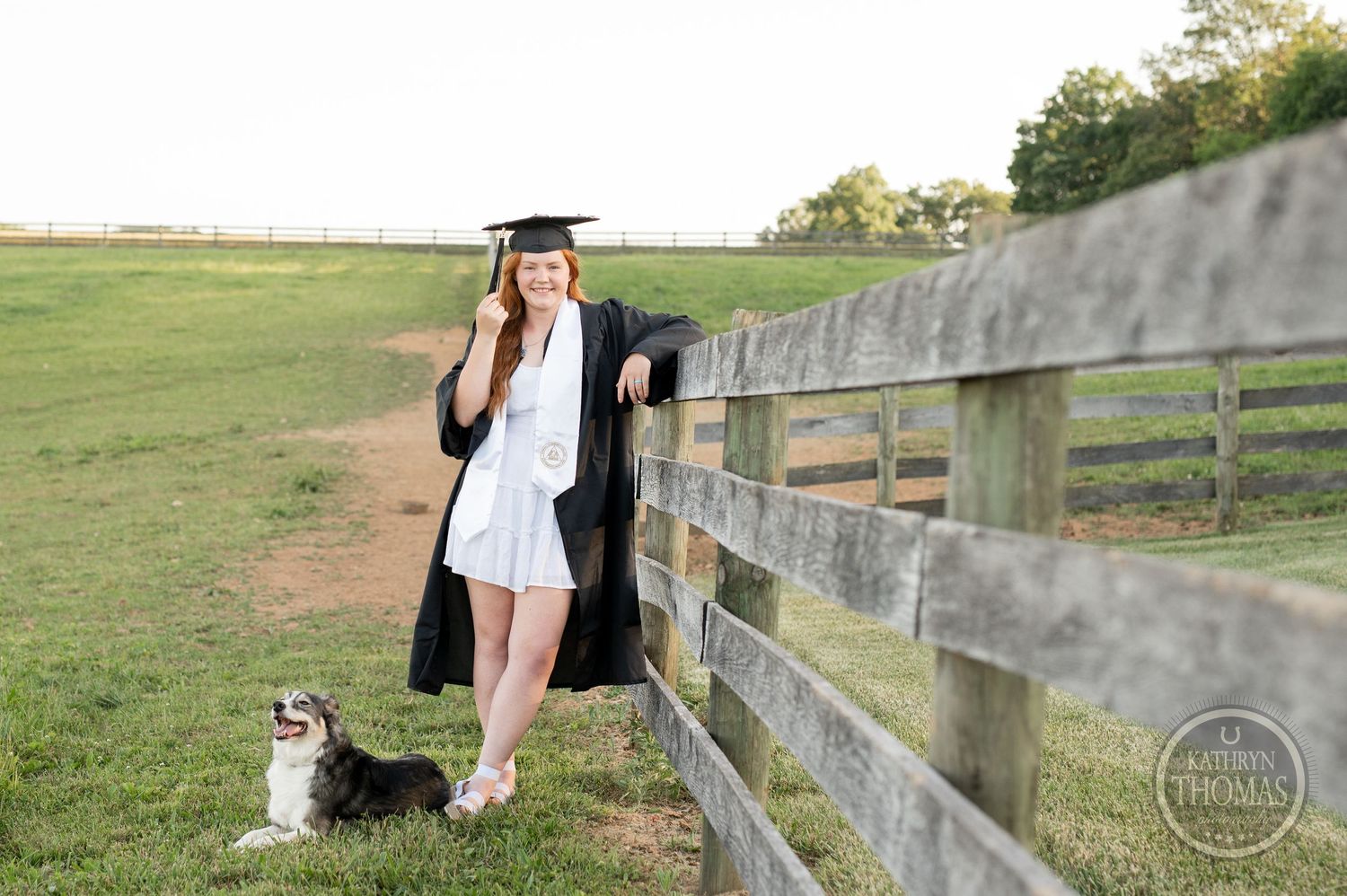 Graduate in cap and gown stands by wooden fence with small dog in scenic field setting.
