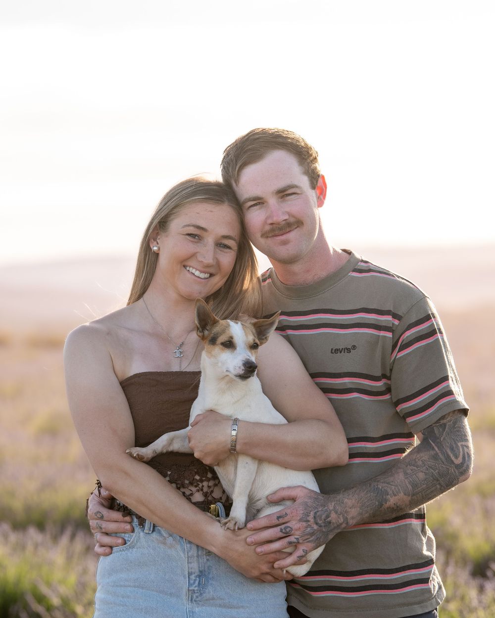 A couple embraces with their small Jack Russell terrier dog during a sunset photoshoot in a natural outdoor setting.