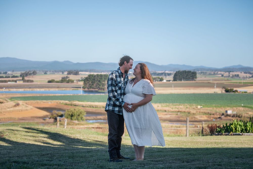 Maternity couple standing together with rural landscape and mountains in background.