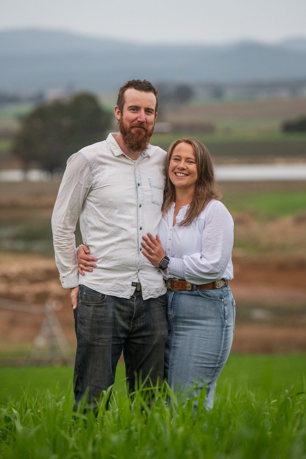 A couple shares a tender moment while standing in a lush green field with rolling hills in the background.