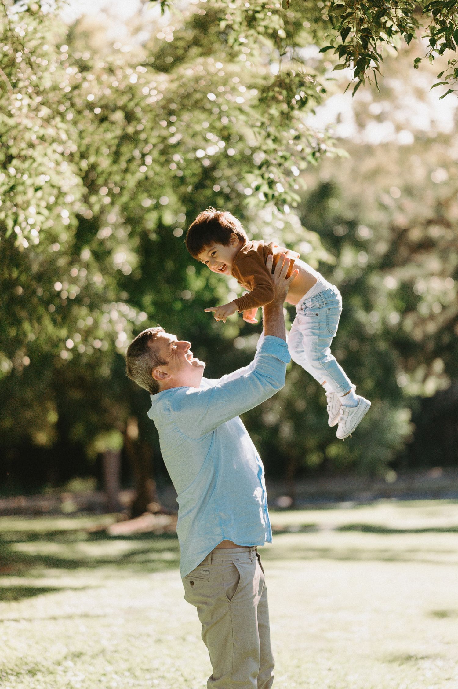 Parent lifting child playfully in sunlit park surrounded by blooming trees.