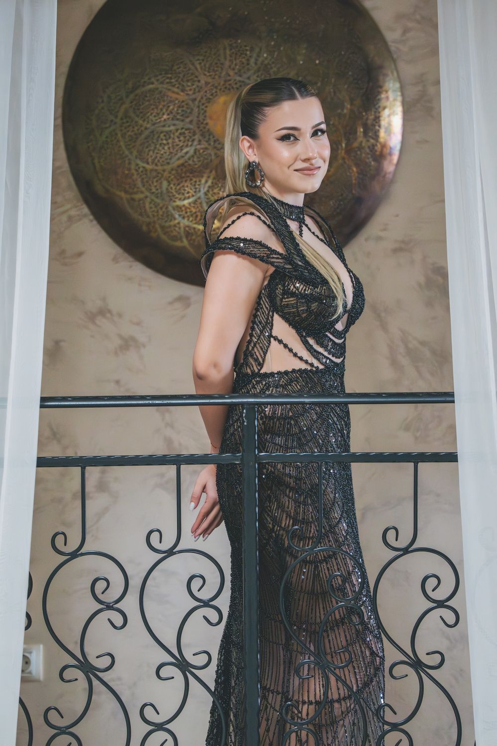 Elegant woman in black lace evening gown poses by decorative railing with ornate metalwork and golden circular wall decor.