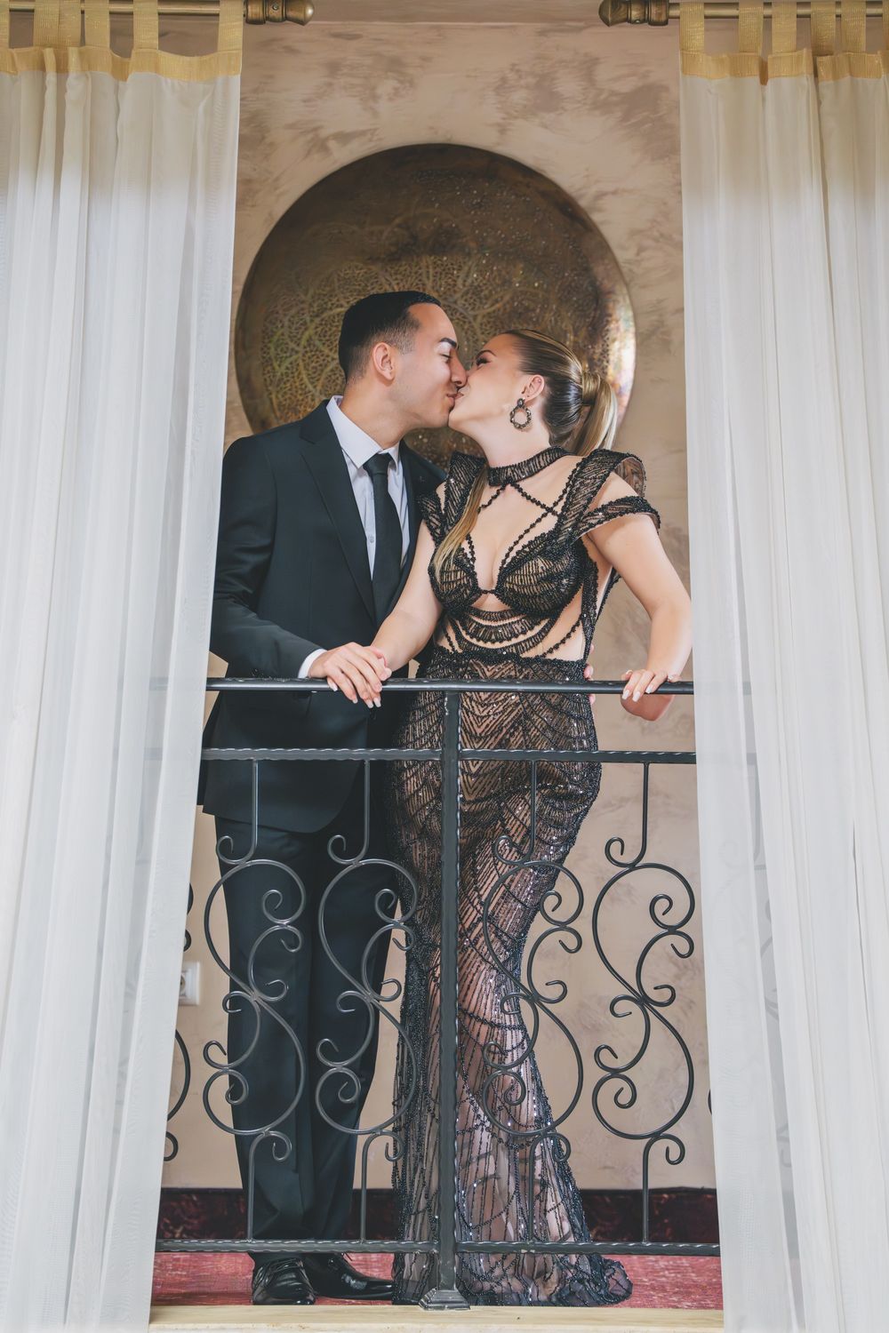 A romantic couple sharing a kiss on a balcony with ornate railings, the woman wearing a black lace gown and man in suit.