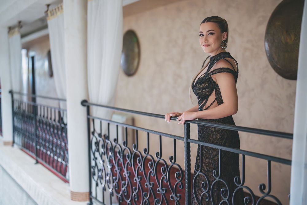 Figure in an elegant black dress poses on a balcony with ornate metal railing against a cream-colored wall.