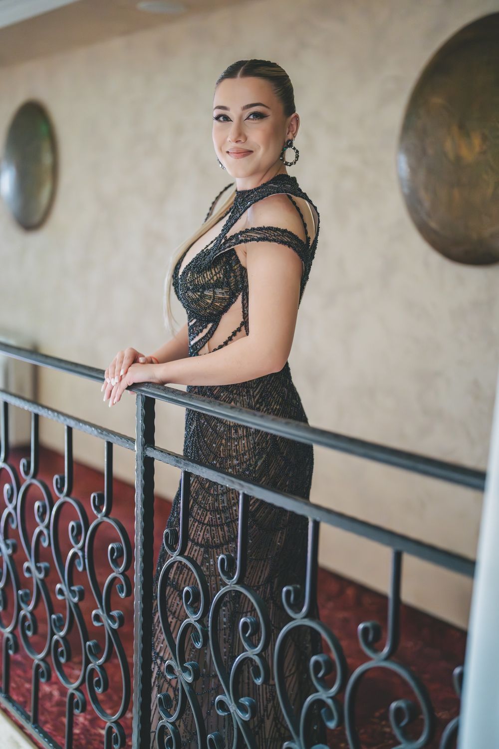 Elegant figure in a shimmering black evening gown poses gracefully against an ornate wrought iron railing in a formal setting.