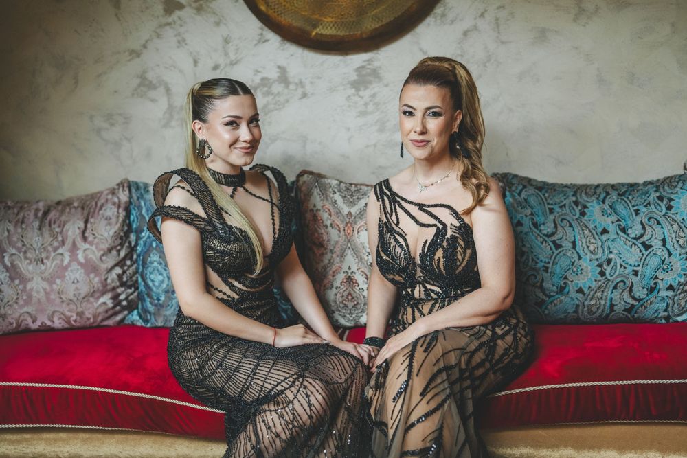 Two people in matching black lace gowns seated on a teal vintage sofa under decorative wall art.
