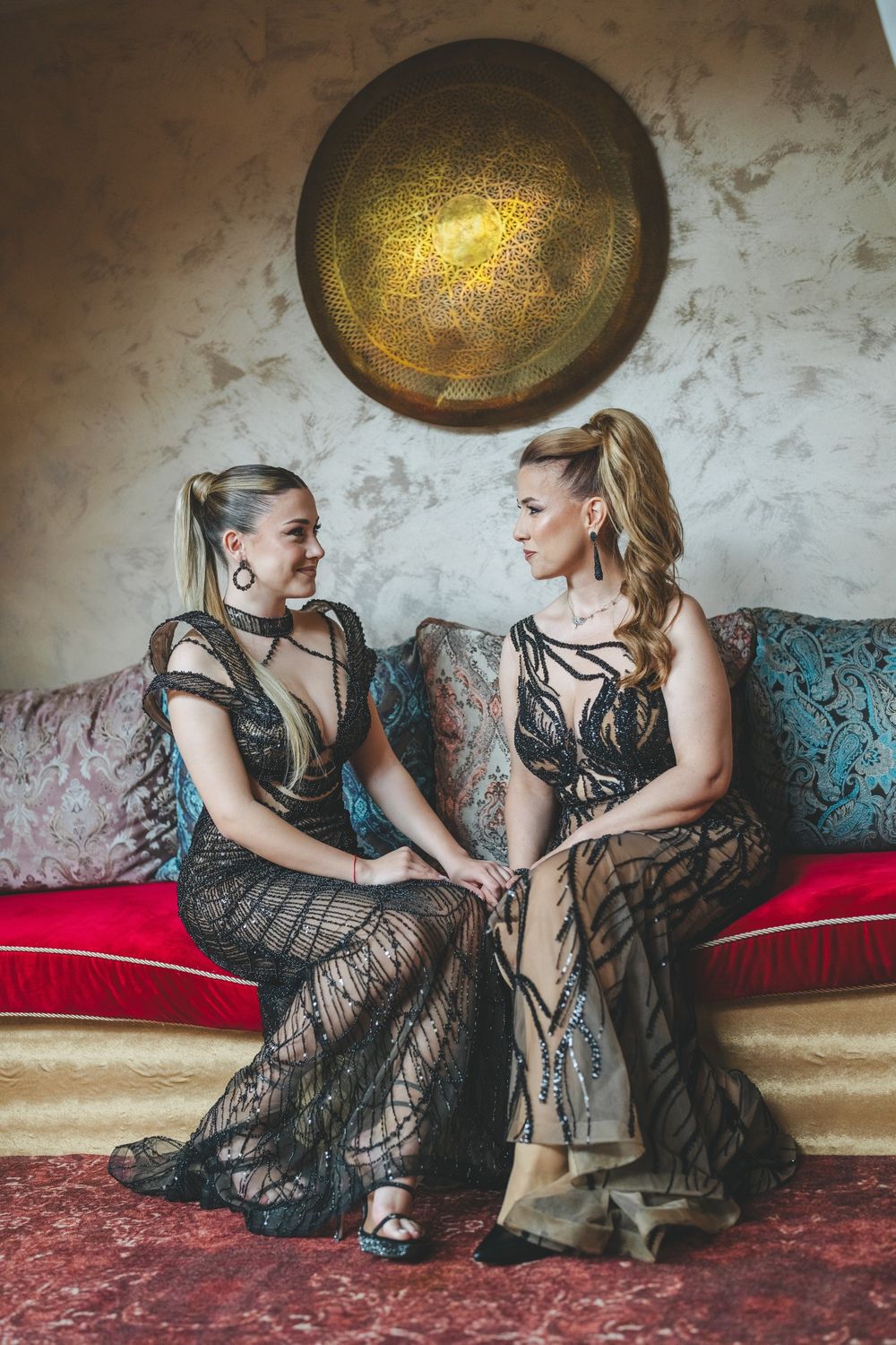 Two people in black lace gowns share a moment on teal sofa under golden wall art.