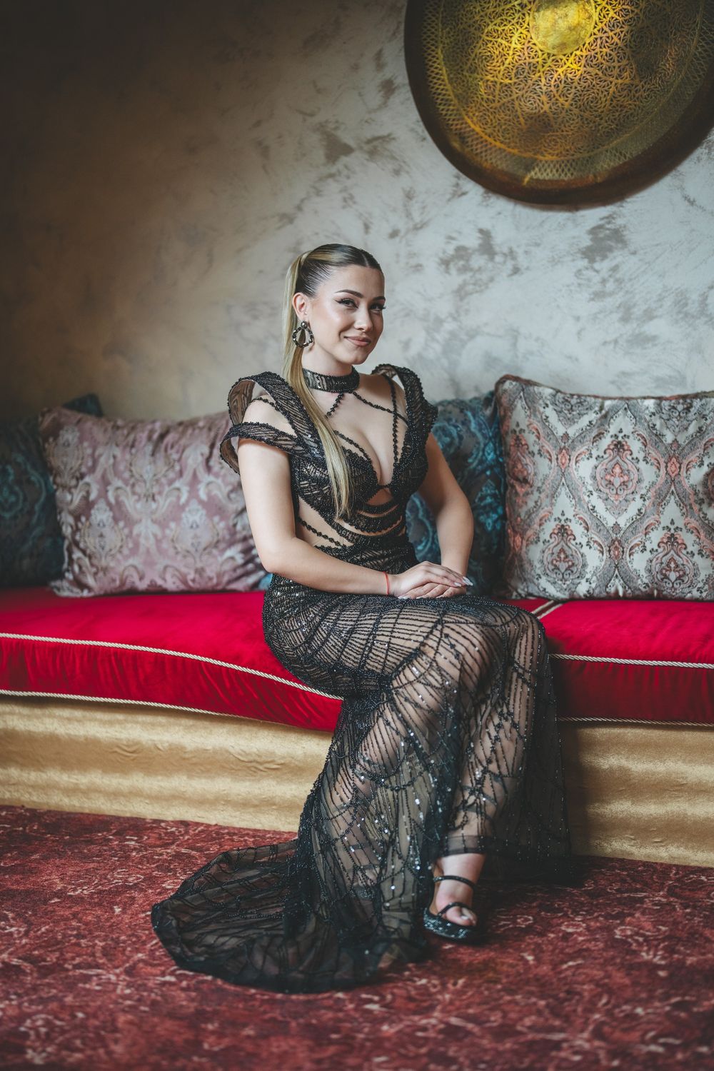 Elegant figure in black lace gown poses on red and teal vintage sofa under golden wall decor.