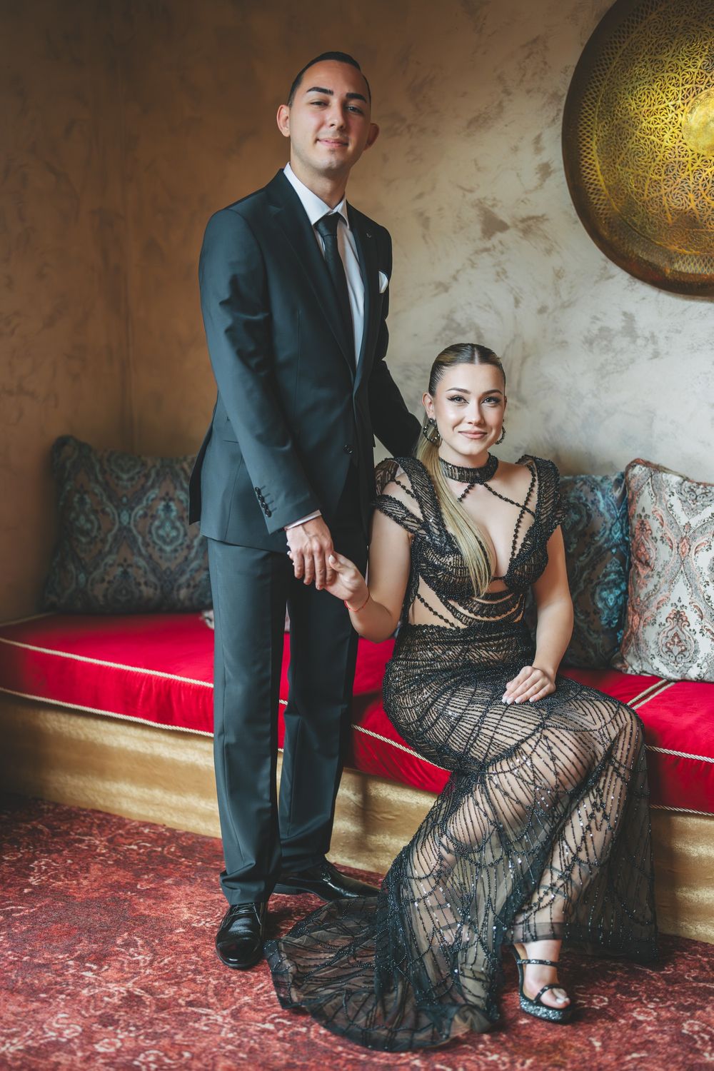 Elegant couple poses in formal attire with a black lace gown and dark suit against a textured wall with decorative globe.