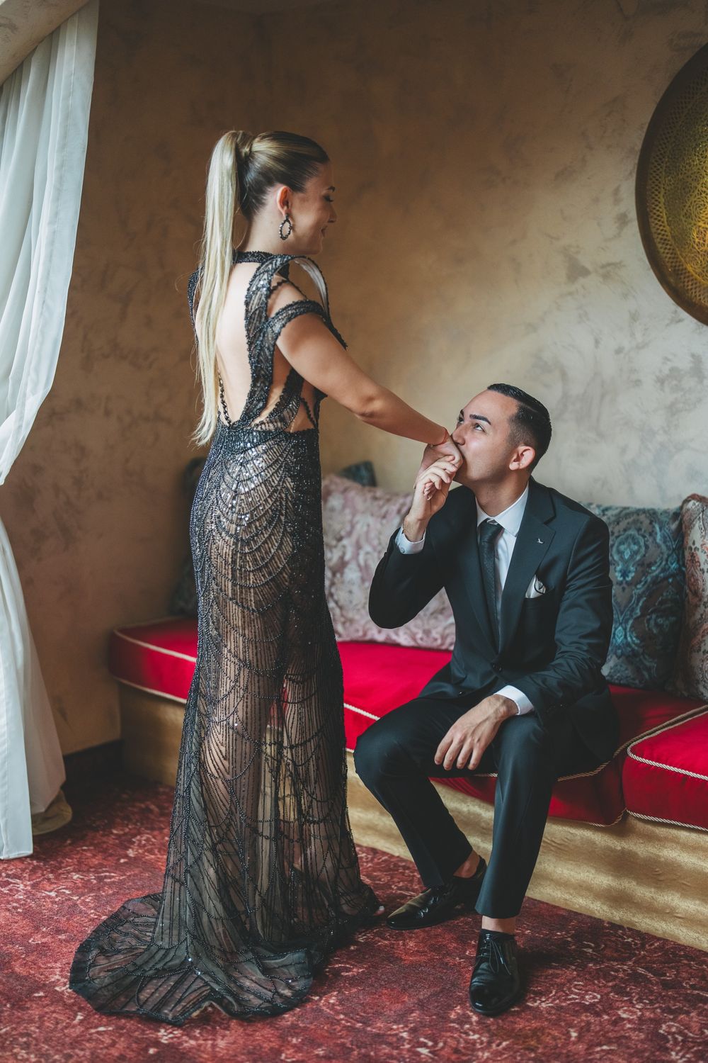 Romantic moment captured as couple interacts on red cushioned bench in ornate setting.