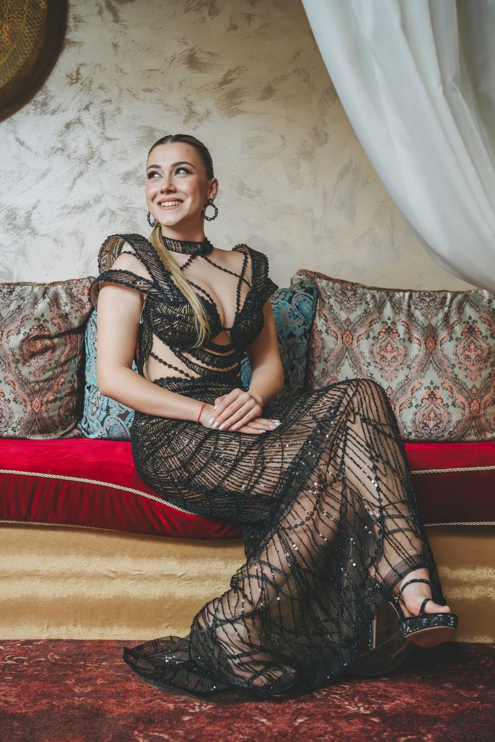 Woman in stunning black lace gown sits elegantly on red cushioned bench in formal setting.