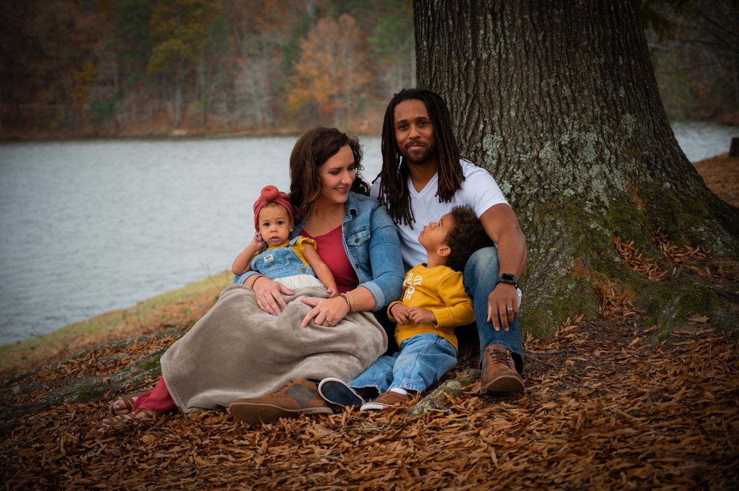 Family relaxing together on autumn leaves near large tree trunk.