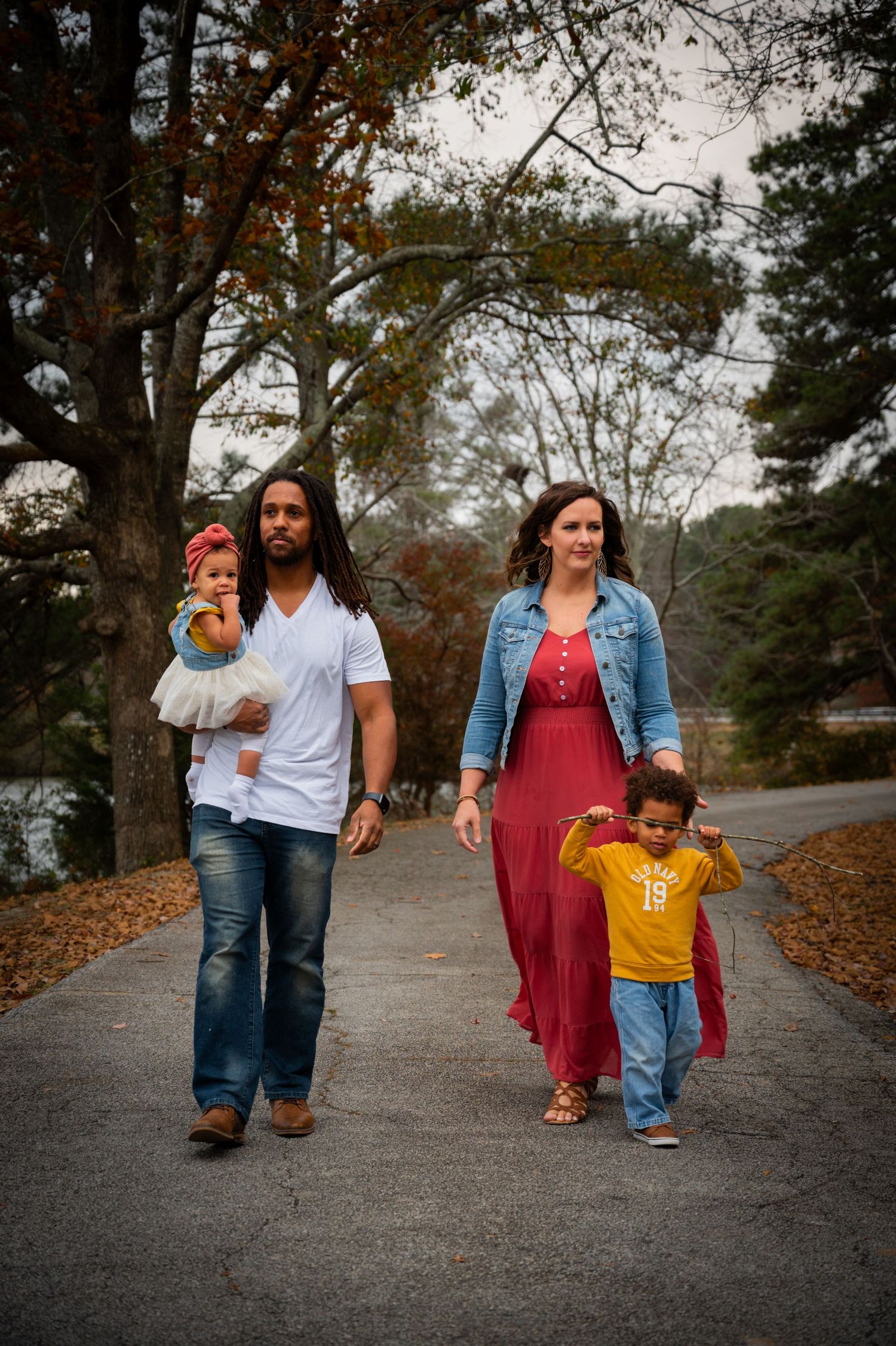 Family strolling down autumn path surrounded by falling leaves and trees.